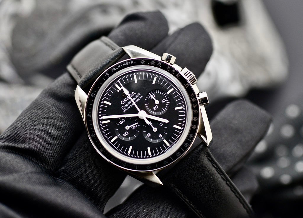 Omega Speedmaster Professional Moonwatch held in a gloved hand, showing black dial, tachymeter bezel, and stainless steel case
